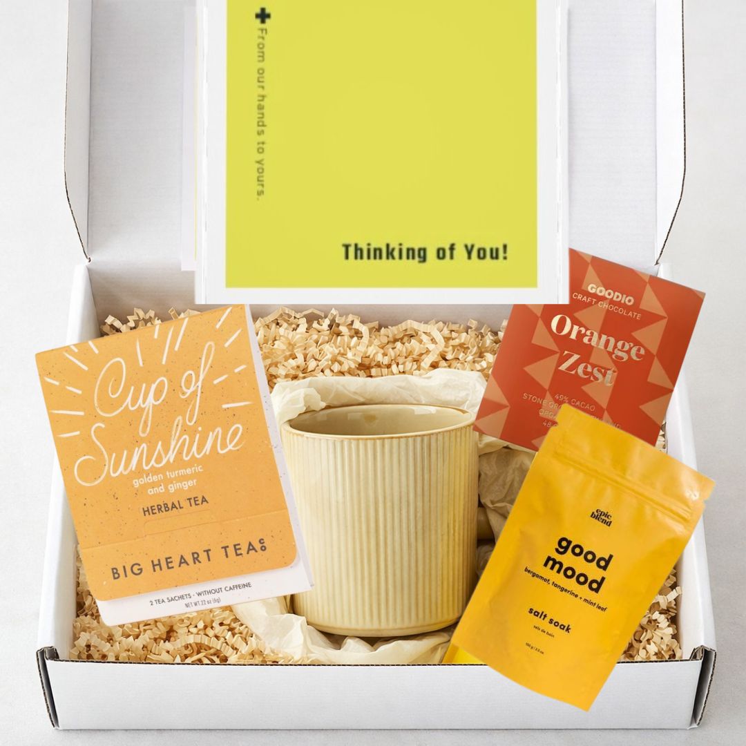 Thinking of you care package with warm-toned mug, sunshine tea, bath salts, and artisan chocolate in a mailer gift box